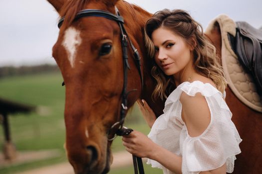 A girl in a white sundress stands next to a brown horse in a field in summer.