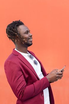 vertical profile portrait of a smiling black businessman in suit typing a message on his smart phone in front of a red background, copy space for text, concept of technology and communication