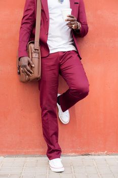 vertical photo of an unrecognizable stylish african businessman dressed in suit with briefcase holding coffee in his hand in front of red background, copy space for text