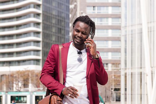 elegant african businessman walks through the city financial downtown talking on the phone with a coffee in his hand, concept of technology and communication, copy space for text