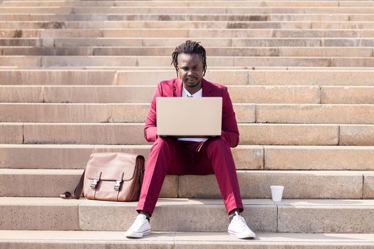 elegant black businessman works with his laptop sitting on a staircase in the city, technology and remote work concept