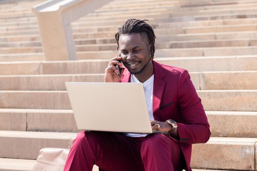 smiling black man works with computer laptop and phone sitting on city stairs, technology and remote work concept
