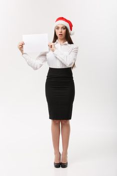 Business Concept - Beautiful young confident business woman with santa hat holding white blank paper with surprising facial expression.