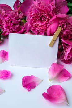 Bright pink peonies on a white background. Summer flower arrangement. Background for a greeting card with paper note. Copy space. The concept of a holiday, birthday, Valentine's Day, gift, declaration of love