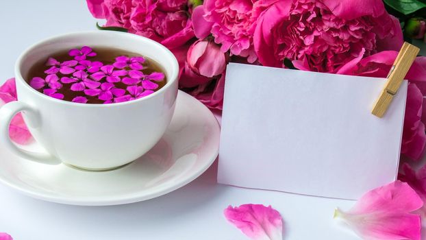 Creative layout with pink peonies flowers and cup of tea on bright table. Paper note. Copy space. Spring Seasonal valentine, woman, mother, 8 march holiday, romance breakfast. Stylish blogger. Greeting card. Invitation. Blog post