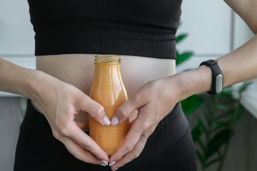 Hand in heart symbol holding smoothie drink detox on fit and slim belly woman. Fitness wellbeing concept. Body care, clean eating, weight loss diet concept. Woman with bowel or reproductive problem. Working out at home. Belly waist exercises