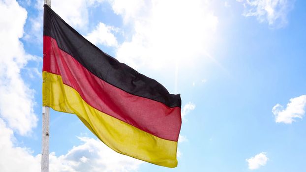 The official flag of Germany waving on a blue sky background. Horizontal banner design, with the German flag hanging on a sunny background with white clouds. Deutschland flag wide banner.