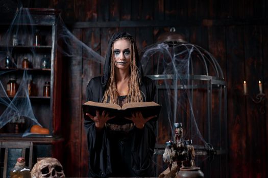 Halloween concept. Witch dressed black hood with dreadlocks standing dark dungeon room use magic book for conjuring magic spell. Female necromancer wizard gothic interior looking camera medium shoot