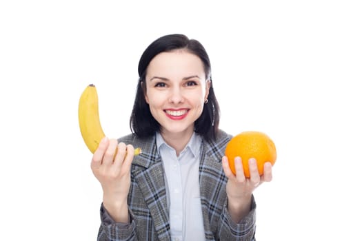 smiling woman in a blue shirt and office suit holding a banana in one hand, an orange in the other, white studio background. High quality photo