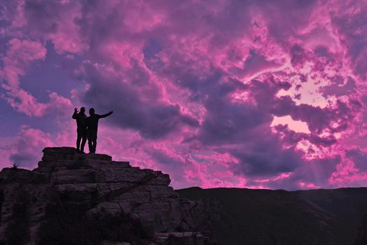 Silhouettes of two men waving their hands stand on a mountain against the backdrop of a neon sky. Achievement concept.