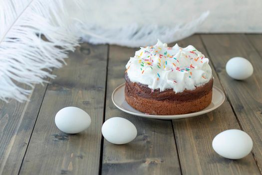 white Easter cake with colored sweet sprinkles near eggs and feathers on a wooden background. Happy easter concept. Soft focus