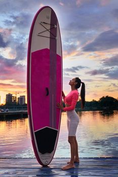 Full length portrait of young woman in activewear standing on wooden pier and holding colorful sup board. Amazing summer sunset behind. Water sport and people concept.