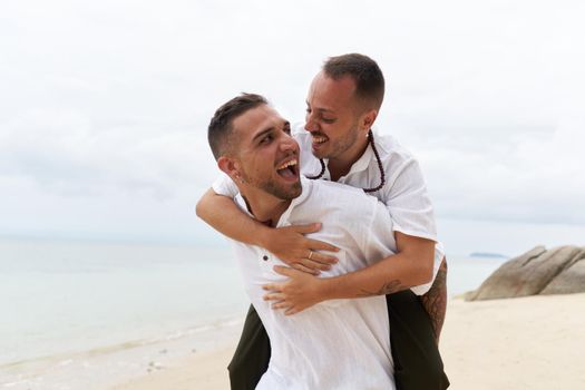 Close up portrait of a smiling man piggybacking his gay partner on a sandy beach