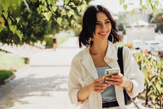 Cheerful Young Woman Holding Smartphone While Walking Outdoors in Park at Summer Sunny Day, Copy Space