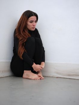 Vertical image of a sad young woman in the corner of a room dressed in black barefoot and on the verge of tears.. Concept of emotions.