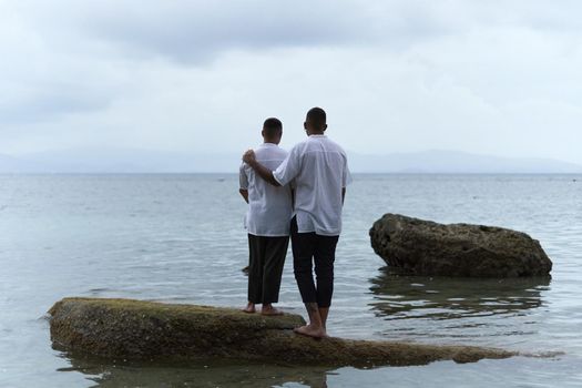 Back of a gay couple standing on a rock looking out to the horizon of the ocean in the evening