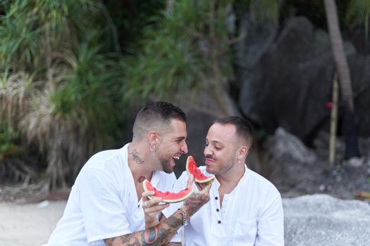 Portrait of a modern gay couple laughing while feeding each other with watermelon on a beach