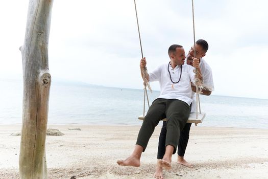 Gay man swinging his partner sitting on a swing in the middle of a tropical beach in Thailand