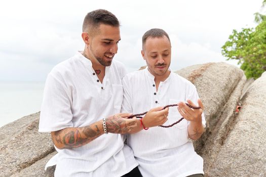 Homosexual couple in white clothes looking at a necklace they are holding in their hands in a beach