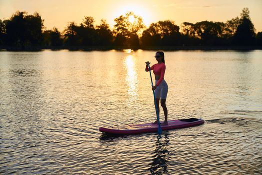 Active young woman in sport clothes and sunglasses spending evening time for practising in sup boarding. Concept of people, leisure and water sport.