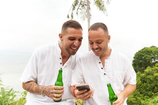 Hsappy caucasian gay couple in white using a mobile while holding beer next to a tropical beach
