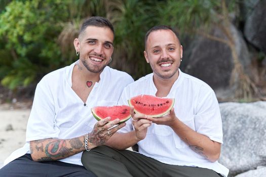 Gay couple looking at camera while showing a piece of watermelon and sitting on a beach