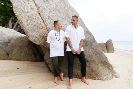 Gay couple leaning on a rock formation on a beach dressed in white and black casual clothes
