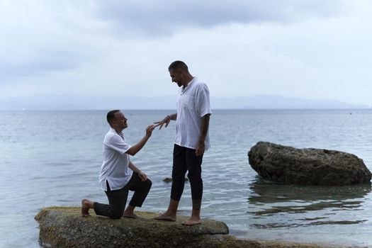 Gay man on his knees asking his partner to marry him in the middle of rocks in the sea