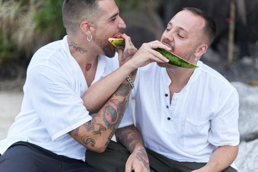 Playful gay couple feeding each other with watermelon on a beach