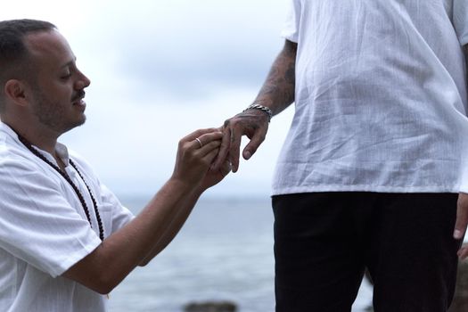 Gay man on his knees asking his partner to marry as he fits the ring on his finger next to the sea