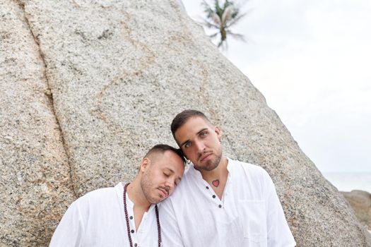 Gay man leaning his head on the shoulder of his couple while standing next to a rock in the beach