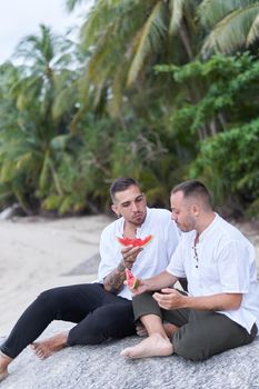 Vertical photo of a gay couple eating watermelon on a tropical and sandy beach in Thailand