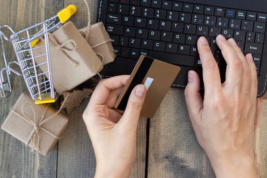 close-up of female hands holding a credit card against the background of keyboard and small basket with gifts. the concept of online shopping, gift shopping. top view