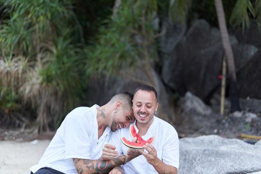 Gay man laughing as he leans on his partner while feeding each other with watermelon on a beach