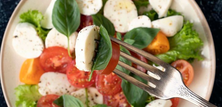 banner with close up mozzarella, tomatoes basil leaf on a fork on a background of vegetable salad with cheese. Top view. Soft focus