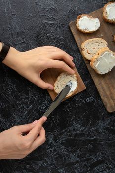 the process of making sandwiches or snacks. female hands smear cream cheese on bread with a knife on a black texture background. Soft focus. top view . flat lay