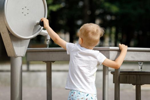 A little boy, wearing shorts and a white T-shirt, plays with water from a tap, he turns a handle and pumps water.