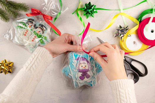 female hands in a white sweater are packing Christmas gifts with gingerbread. gingerbread cookies with snowmen, scissors, wrapping paper, colorful ribbons on the table. flat lay top view