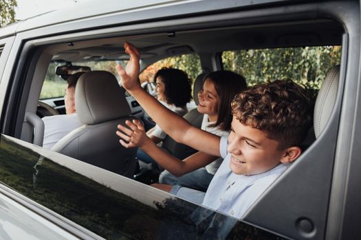 Teenage Boy and Girl Greeting to Someone Through Window with Waving Hands While Sitting Inside Minivan Car, Happy Four Members Family on Weekend Road Trip