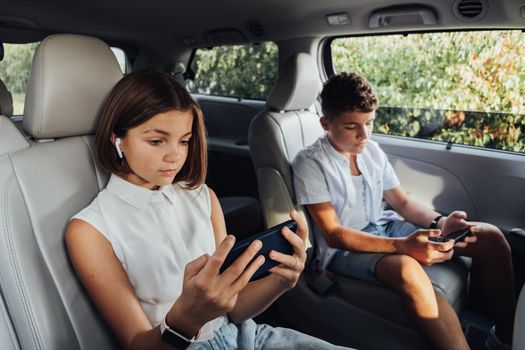 Young Caucasian Girl and Her Teenage Brother Focused on Smartphones While Sitting in Minivan, Children Playing Games on Weekend Family Road Trip by Car