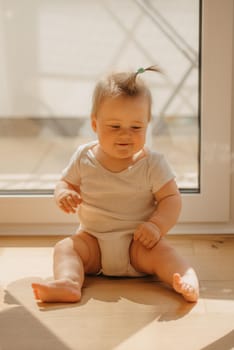 A 7-month girl is sitting near a balcony door in a bodysuit at home. A cute infant is staring to the side in the sunlight.