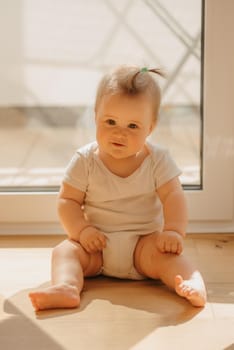 A kind 7-month girl is sitting near a balcony door in a bodysuit at home. A cute infant is waiting for her parents in the sunlight.