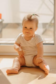 A kind 7-month girl is sitting near a balcony door in a bodysuit at home. A cute infant is smiling in the sunlight.