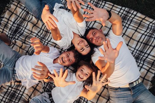 Top Down View of Four Members Family Having Picnic Outdoors at Sunset, Caucasian Mother and Father with Two Teenage Children Laying on Plaid and Raising Hands Up to Camera