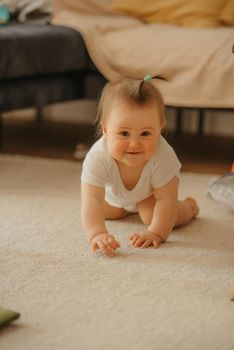 A 7-month girl is crawling near a sofa and smiling at home. An infant in a bodysuit in the sunlight.