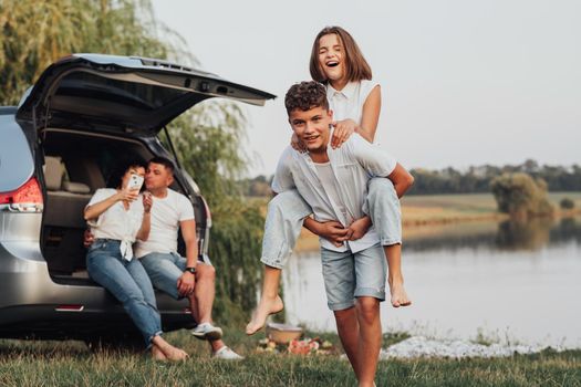 Two Happy Teenage Children Playing by Lake While Their Parents Sitting in Trunk of the Minivan Car, Four Caucasian Members Family Enjoying Road Trip Outdoors