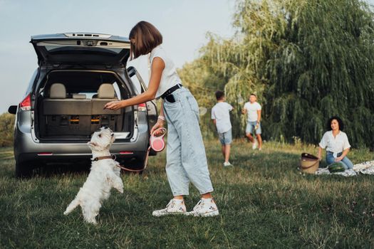 Teenage Girl Playing with West Highland White Terrier Dog on Background of Her Family Having Fun Time Outdoors on Road Trip by Minivan Car