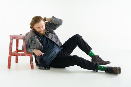 a handsome brutal bearded and long-haired man sits on the floor leaning on a chair on a white background, he is dressed in a denim jacket and pants with bright green socks, dressed like a hipster. High quality photo