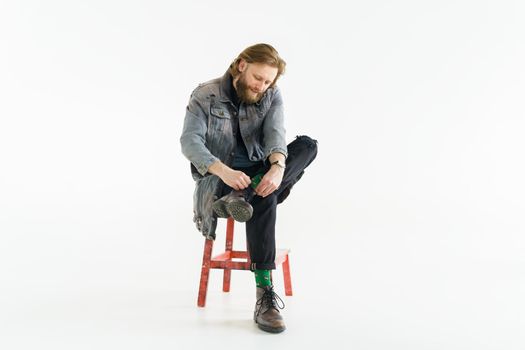 a handsome brutal bearded and long-haired man is sitting on chair on a white background, he is dressed in a denim jacket and pants with bright green socks, dressed like a hipster. High quality photo