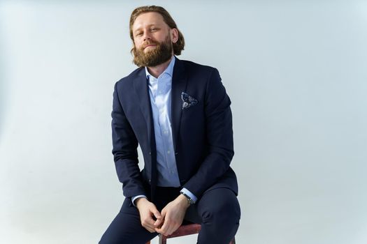 a handsome brutal bearded and long-haired man, a stylish businessman sitting on a chair with a white background. High quality photo